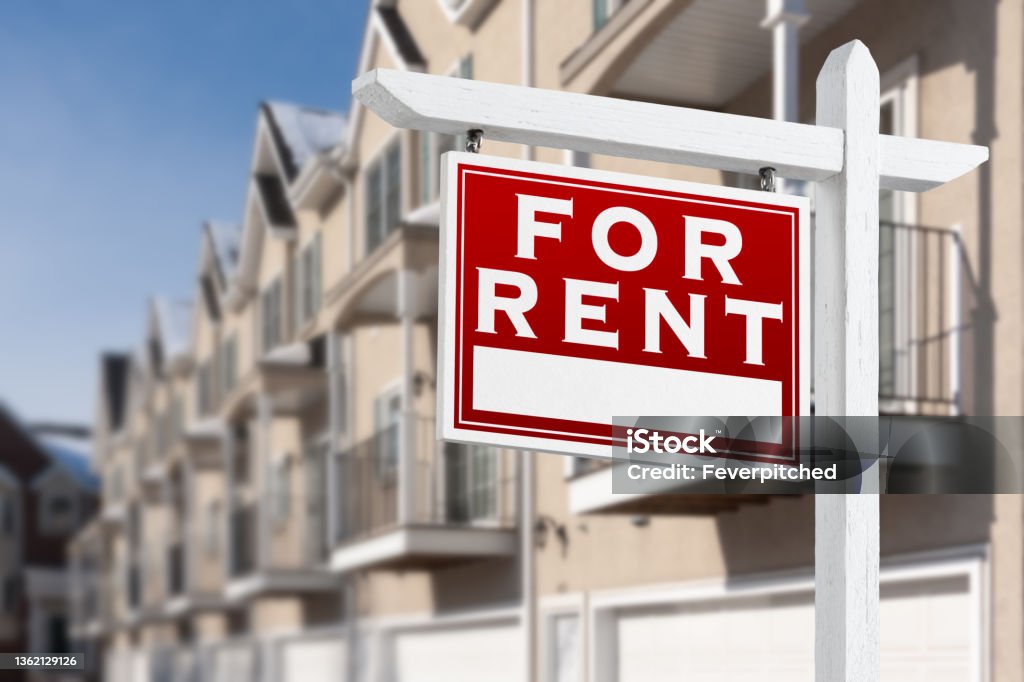 For Rent Real Estate Sign In Front of a Row of Apartment Condominiums Balconies and Garage Doors.